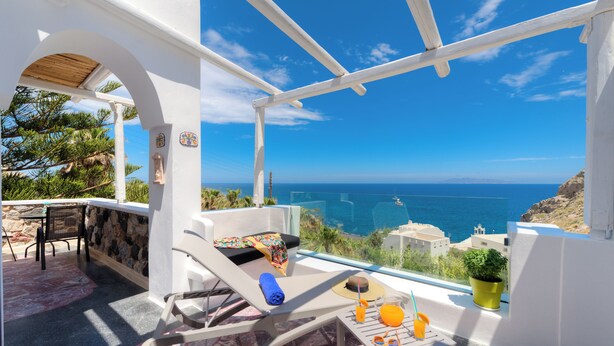A balcony with a view of the sea, a table with a hat, sunglasses, and a drink, and a chair with a towel. - Moschatos Beach House (Santorini)