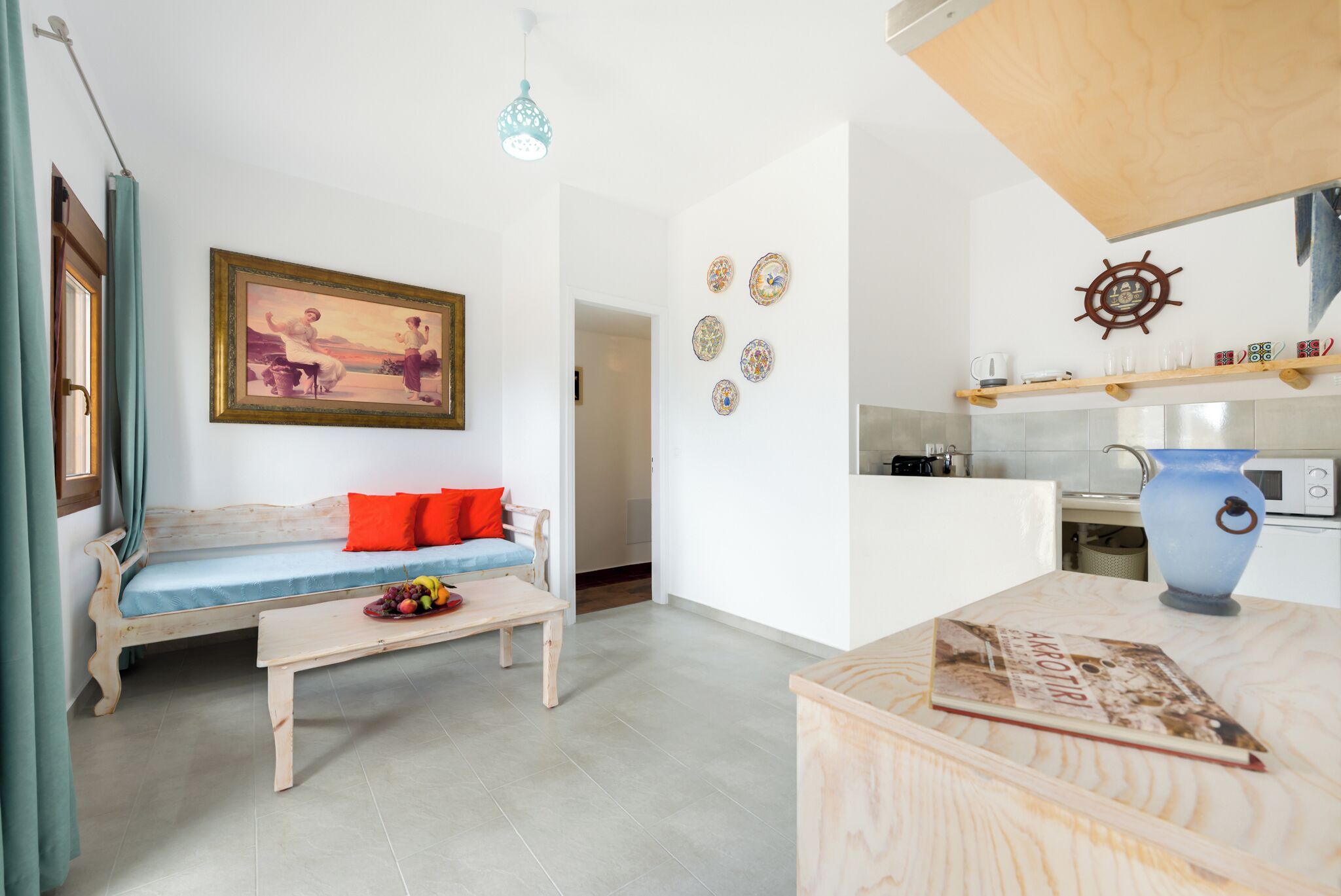 Un salon avec un canapé, une table basse sur laquelle se trouvent des fruits, un tableau, et un coin cuisine avec une cuisinière et un vase bleu.