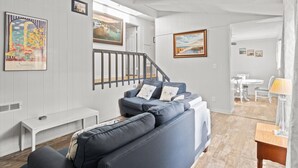 A modern living room with a grey sofa, a white bench, and a staircase leading to another room. - Beachy Getaway • Clean, Private 3BR (Wendell)