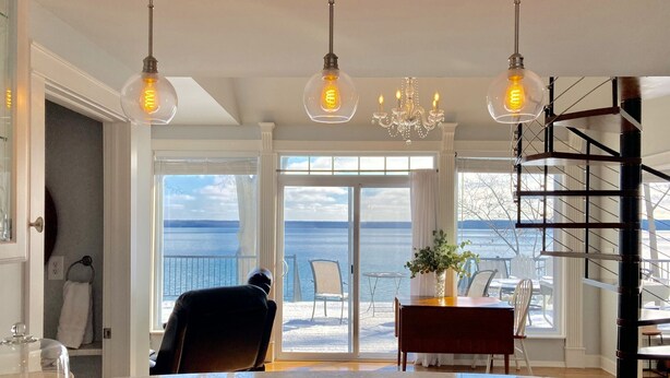 A spacious living room with a large glass door leading to a balcony overlooking a body of water. There are three hanging light fixtures and a chandelier. - Smith's Lake House 4 bedroom, enjoy spectacular sunrises, 2 bathroom, built 2021 (Penn Yan)