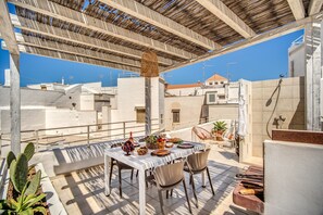 Un área de comedor al aire libre con una mesa preparada para cuatro personas, rodeada de sillas, bajo una pérgola de madera.