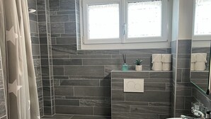 Un baño moderno con bañera, inodoro y lavamanos, con azulejos oscuros y una ventana sobre el inodoro.