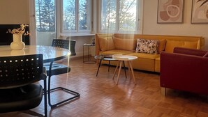 Una sala de estar moderna con suelo de parqué de madera, un sofá amarillo, un sillón rojo, una silla negra, una mesita blanca pequeña y una lámpara colgante original.
