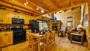 Interior acolhedor de uma cabana com pisos de madeira, sala de jantar com cadeiras, lareira e cozinha com armários e eletrodomésticos de madeira.