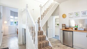 A staircase with white balusters and striped carpet leading to a second floor. A kitchen with a stainless steel refrigerator and a countertop with various items is visible.