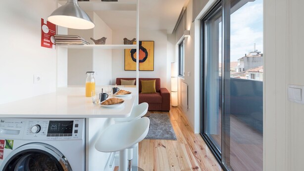 A modern kitchen with a washing machine, dining area with a table and chairs, a red sofa, and a large window with a city view. - Gorgeous Modern Flat w/ Balcony by LovelyStay (Porto)