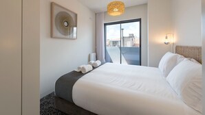 A hotel room with a large bed, a view of the city through a sliding glass door, a framed artwork on the wall, and a ceiling light fixture. - Gorgeous Modern Flat w/ Balcony by LovelyStay (Porto)