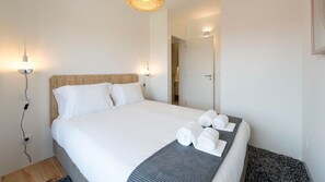 Cama bem arrumada com roupa branca e toalhas dobradas, cabeceira de tecido e abajur.