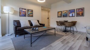 Un salon moderne avec un canapé en cuir marron, une table basse et un coin repas avec des chaises en osier. Des affiches de voyage encadrées ornent le mur.
