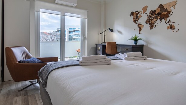 Ein modernes Hotelzimmer mit einem großen Bett, einer Sitzecke mit einem Sessel, einem Schreibtisch, einer Weltkarte an der Wand und Blick auf die Stadt durch das Fenster.
