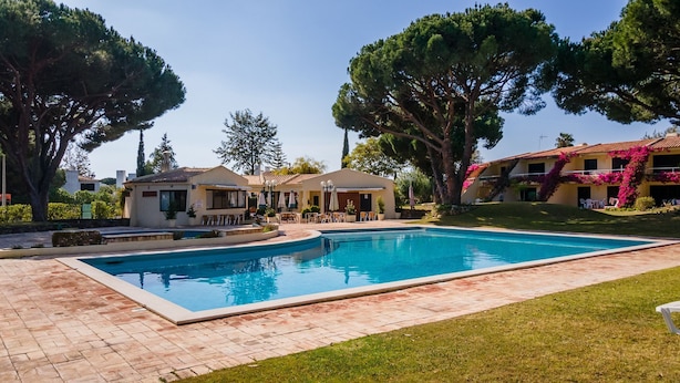 A swimming pool with a tiled deck, surrounded by trees and buildings with outdoor seating. - Gorgeous 2BDR W/Patio & Pool by LovelyStay (Vilamoura)