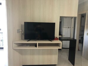 Ein modernes Wohnzimmer mit Flachbildfernseher auf einem Holztisch, einer Küchenzeile mit Kühlschrank und einer Tür zu einem weiteren Raum.
