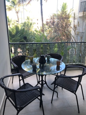 En balkon med et glasbord og fire fletstole, med udsigt over en frodig have med palmer.