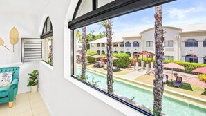 A room with a large window overlooking a pool and a courtyard with palm trees and outdoor seating. - Tropical Escape at Mango Lagoon Resort – Spacious 2-Bedroom Apartment with Pools, Palm Cove Charm, and Easy Esplanade Access (Palm Cove)
