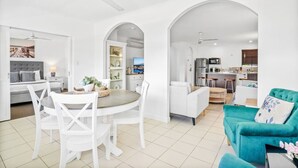 A spacious living area with a dining table, white chairs, and a teal sofa. There is a kitchen area with modern appliances and bar stools. - Tropical Escape at Mango Lagoon Resort – Spacious 2-Bedroom Apartment with Pools, Palm Cove Charm, and Easy Esplanade Access (Palm Cove)