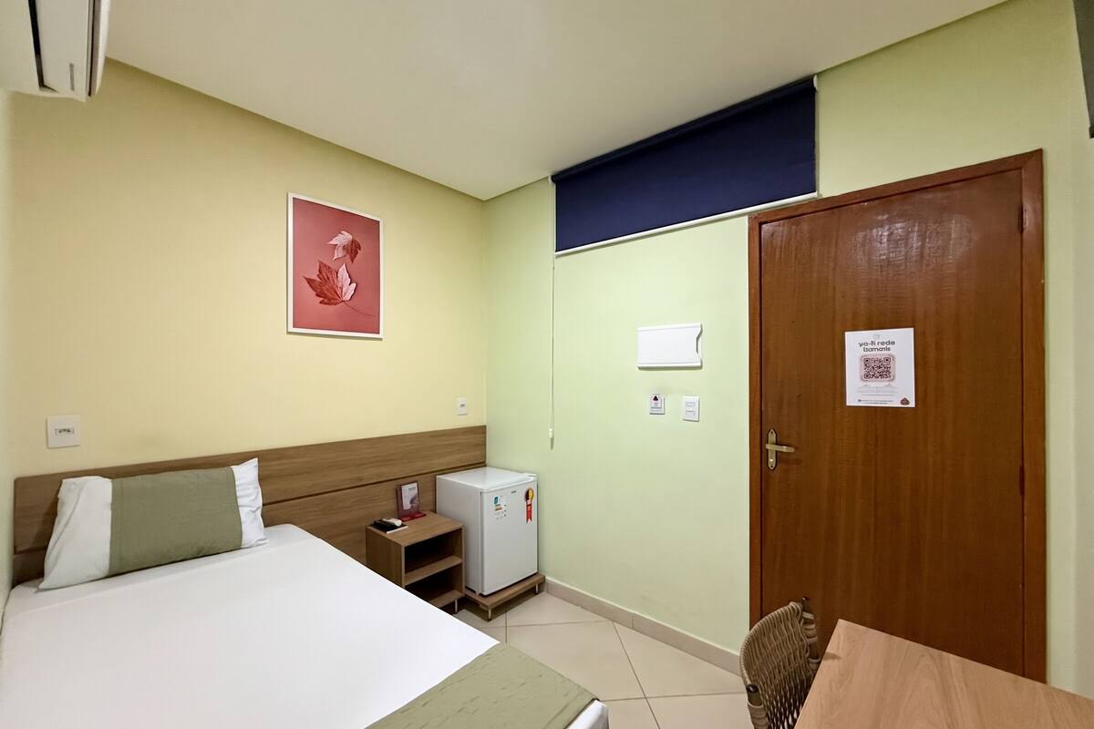Hotelkamer met een bed, een klein tafeltje, een stoel, een koelkast, een houten deur en een wandairconditioner.