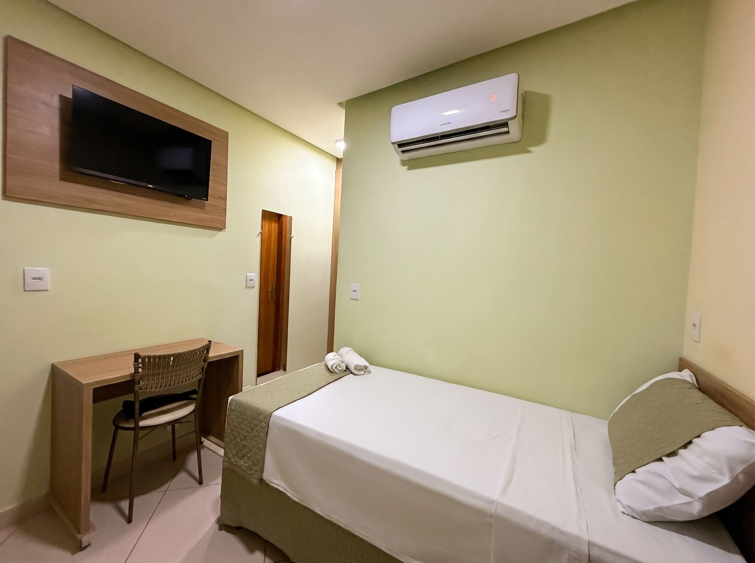 A hotel room with a bed, a desk, a chair, an air conditioning unit, and a television mounted on the wall.