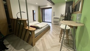 A compact room with a bed, a small desk, and a chair. There is a window with curtains, a mirror, and a wall-mounted air conditioner. - Le Domaine du Verger, Chambres D'hôtes (Osenbach)