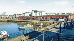 A waterfront marina with boats docked, a red building with arches, and a modern building in the background.