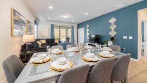 Un comedor con una mesa dispuesta para seis personas, un sofá, un televisor y espejos decorativos en la pared.