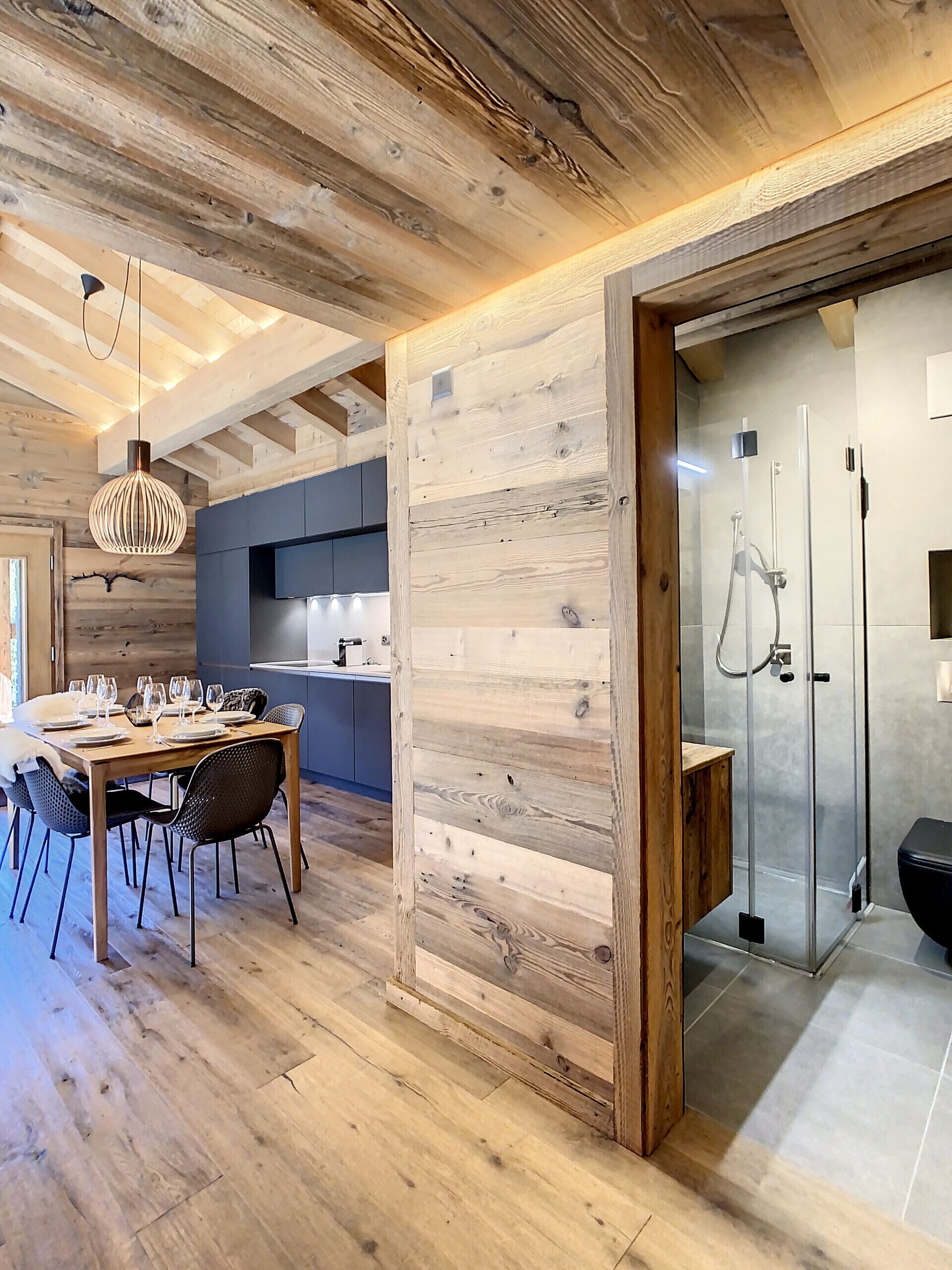 A modern kitchen with wooden accents, a dining area with a wooden table and chairs, and a glass-enclosed shower area.