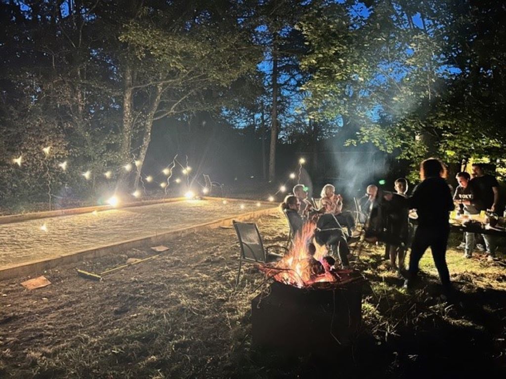 Reunión al aire libre con una fogata, rodeada de árboles y luces colgantes.