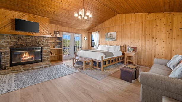Un acogedor interior de cabaña de madera con chimenea de piedra, una cama, un televisor montado en la pared y techo de madera.