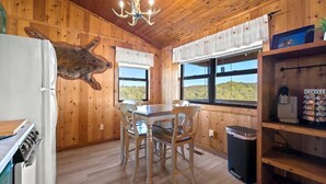 Interior de madera con mesa de comedor, sillas, una lámpara de araña, refrigerador, cafetera y vistas al exterior.