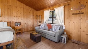 Una habitación acogedora tipo cabaña de madera con una cama, un sofá, una mesita y una ventana con cortinas.