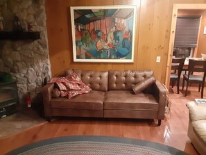 Een bruine leren bank met een plaid, een stenen muur, een schilderij en een eethoek met stoelen en een tafel.