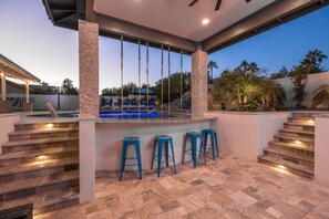 Ein überdachter Außenbereich mit Pool, Steintreppe und blauen Barhocker.