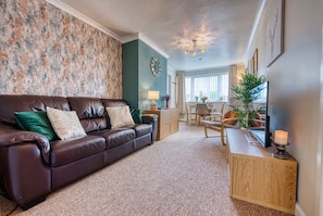 A living room with a brown leather sofa, a wooden side table, and a dining area with a chair and table. - Heather Apt | 3 bedrooms | Sleeps 6 | Southam Centre |Close to HS2 Coventry, Rugby & M40 | Inspire Homes (Southam)