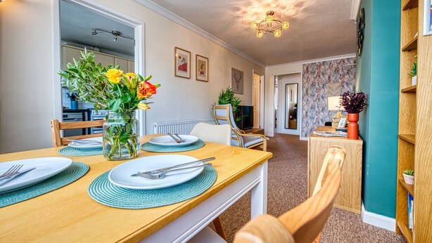 A dining area with a wooden table set for two, a vase with flowers, and a view into the kitchen.