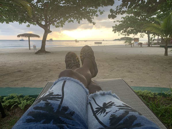 A beach scene with a person relaxing on a lounge chair, a tree, and boats in the background.