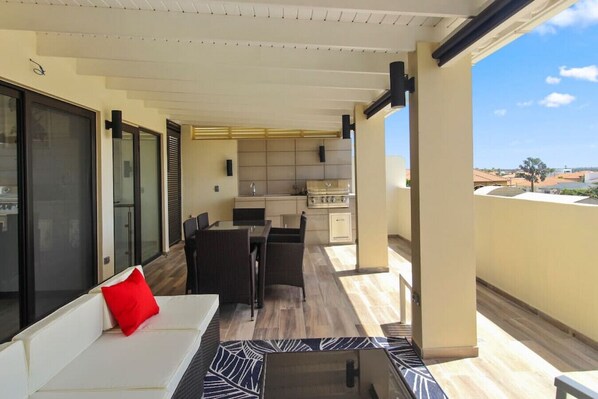 Een balkon met een eethoek, een bank met een rood kussen, en een keuken op de achtergrond.