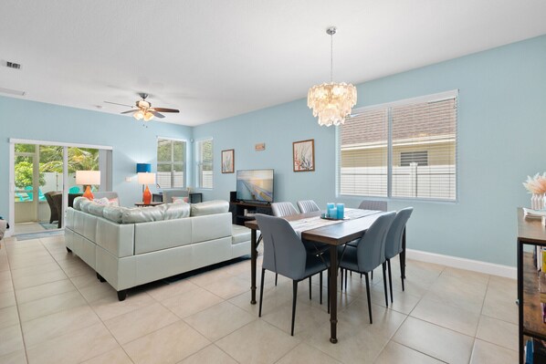 Uma sala de estar espaçosa com parede azul clara, um sofá branco, uma mesa de jantar com cadeiras, um ventilador de teto e um candelabro.