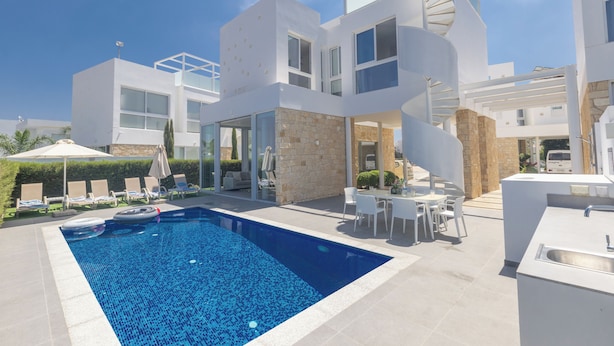 Un área de piscina exterior moderna con una piscina azul cristalina, una escalera espiral y una cocina exterior blanca.