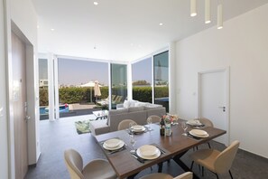 A modern dining area with a table set for four, large windows, and a view of a swimming pool and greenery. - Villa Bailly, Brand New, Elegant 3BDR Protaras Villa with Private Pool (Protaras)
