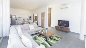 Una sala moderna con un sofá, una mesa de centro y un televisor. Se observa una cocina al fondo con armarios y un refrigerador.
