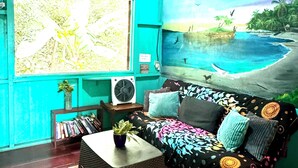 A room with a teal wall, a mural of a beach scene, a sofa with patterned cushions, a small table with a plant, and a bookshelf. - Hidden Jungle Beach House, a Unique Guesthouse in Puerto Viejo de Talamanca!  (Puerto Viejo de Talamanca)