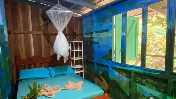 A room with a bed, a bedside table with a plant, a window with a view of greenery, and a mural on the wall. - Hidden Jungle Beach House, a Unique Guesthouse in Puerto Viejo de Talamanca!  (Puerto Viejo de Talamanca)
