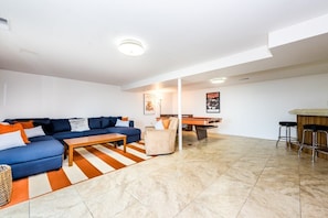 Uma sala de estar moderna com um sofá azul, uma mesa de centro de madeira e um tapete listrado. Há uma mesa de bilhar e um espaço bar ao fundo.