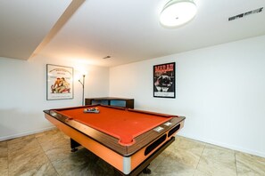 Uma mesa de bilhar em um quarto com paredes brancas, dois cartazes emoldurados e uma luminária no teto.