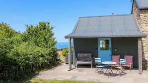 A small, single-story building with a blue door and a metal roof. It has a patio with a bench and two striped chairs. - Dolwylan Barn (Llandysul)
