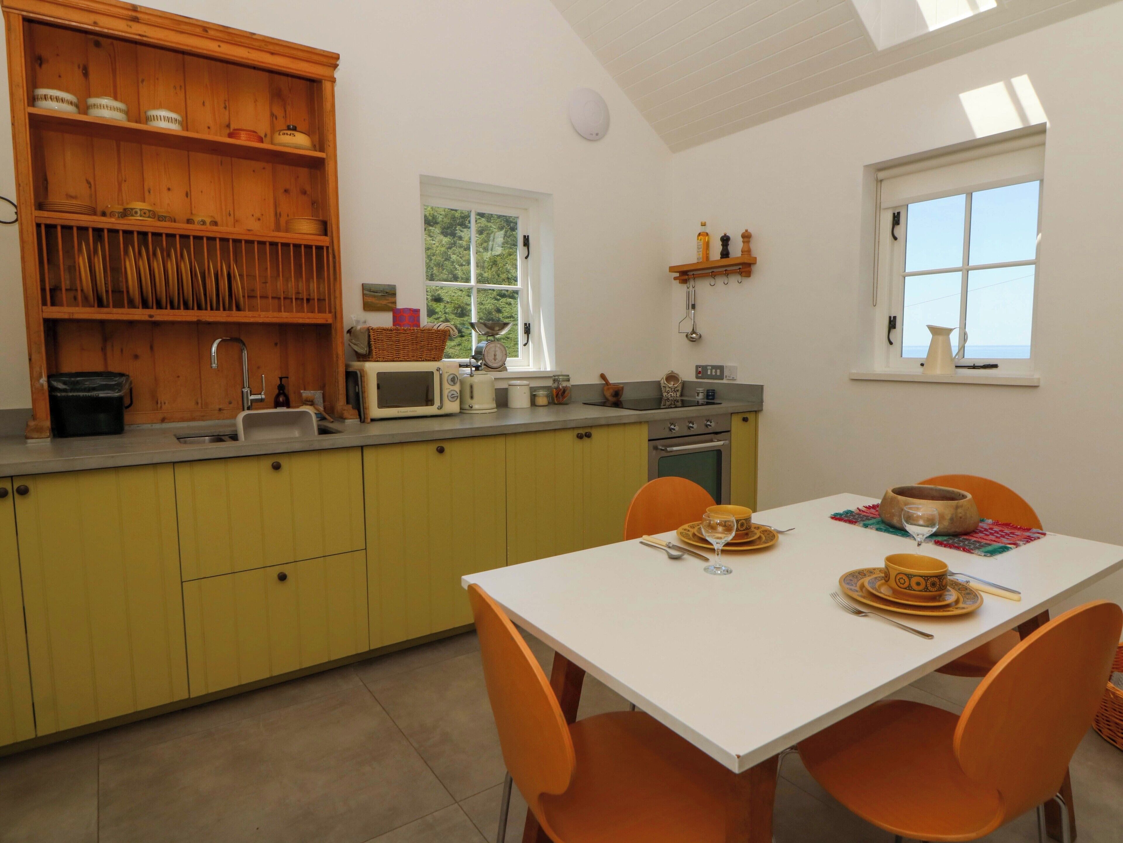 Una cocina con armarios amarillos, encimera blanca y una mesa de comedor naranja con sillas. Se aprecia una ventana con vistas a zonas verdes.
