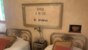 Een slaapkamer met twee bedden, elk met een decoratief kussen en een klein nachtkastje met een vaas. Boven de bedden hangt een lijst met een citaat op de muur.