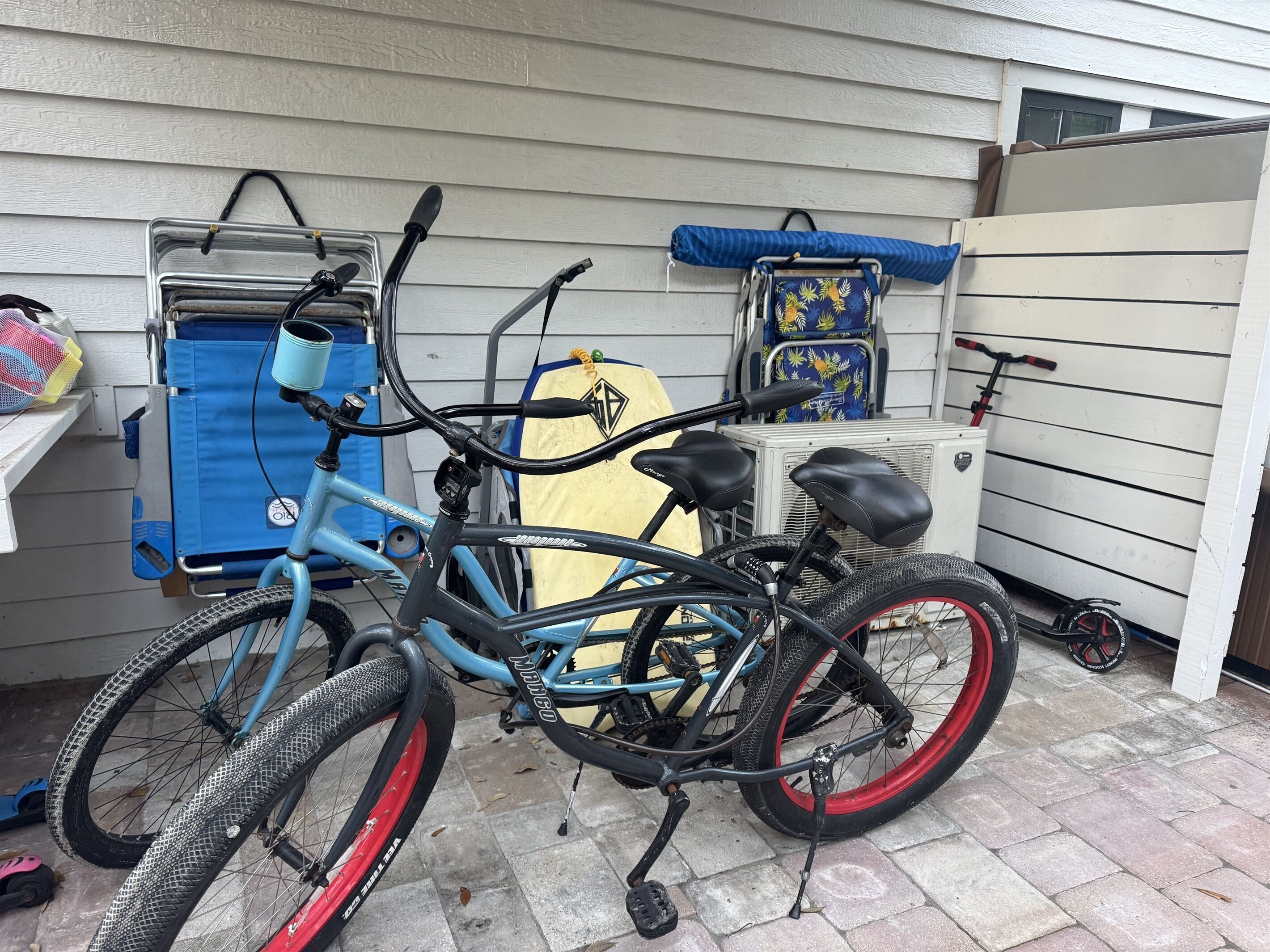 A blue bicycle with a black seat and red rims, parked in a covered outdoor area with a white wall and a blue chair in the background.