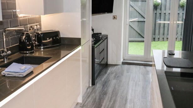 A modern kitchen with a black countertop, stainless steel appliances, and a flat-screen TV mounted above the sink. - Sunnydene (Paignton)