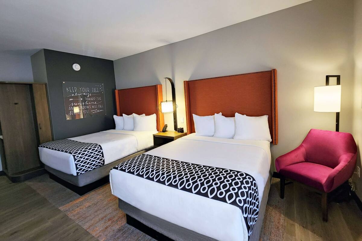 A hotel room with two beds, a red armchair, a wall-mounted light, and a motivational quote on the wall.