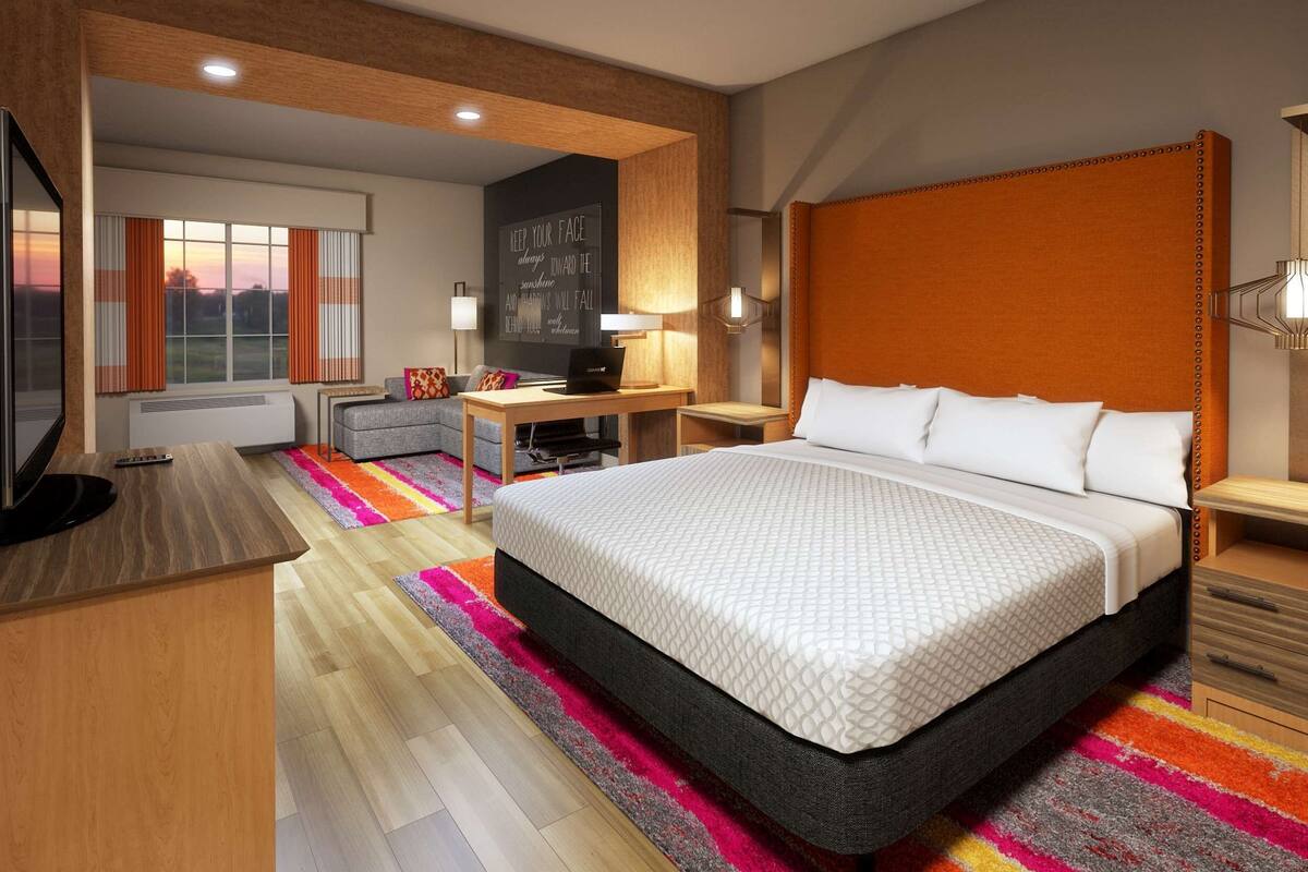 A modern hotel room with a large bed, a desk with a laptop, a TV, and a view of the sunset through the window.